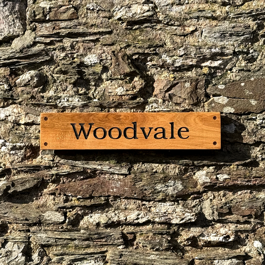 A rectangular wooden sign engraved with the name 'Woodvale' in black lettering, mounted on a rustic stone wall.