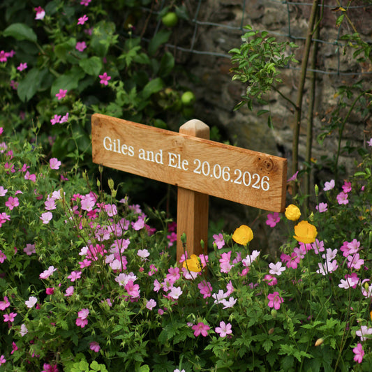 Wooden sign with 'Giles and Ele 20.06.2026' in a garden with flowers and a stone wall.
