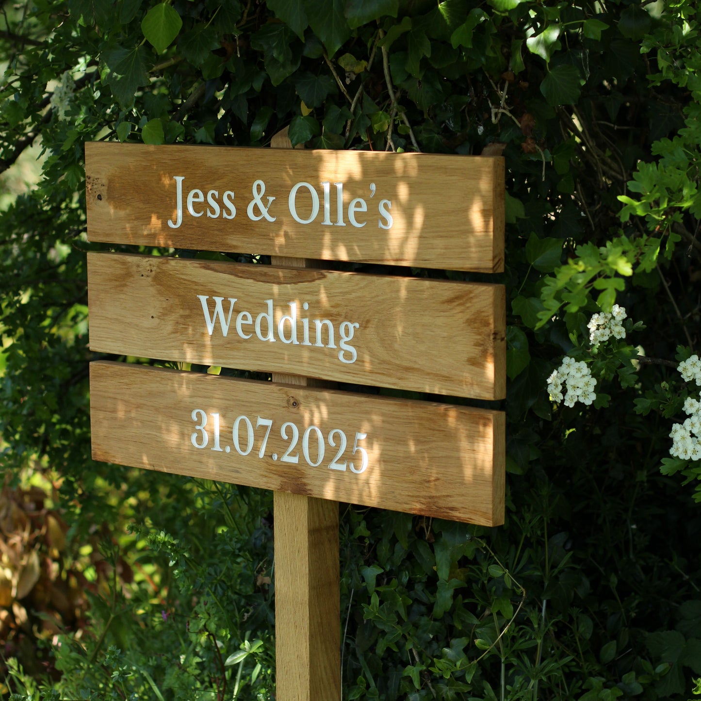 A wooden wedding sign with 'Jess & Olle's Wedding 31.07.2021' engraved on it, set against a backdrop of greenery.