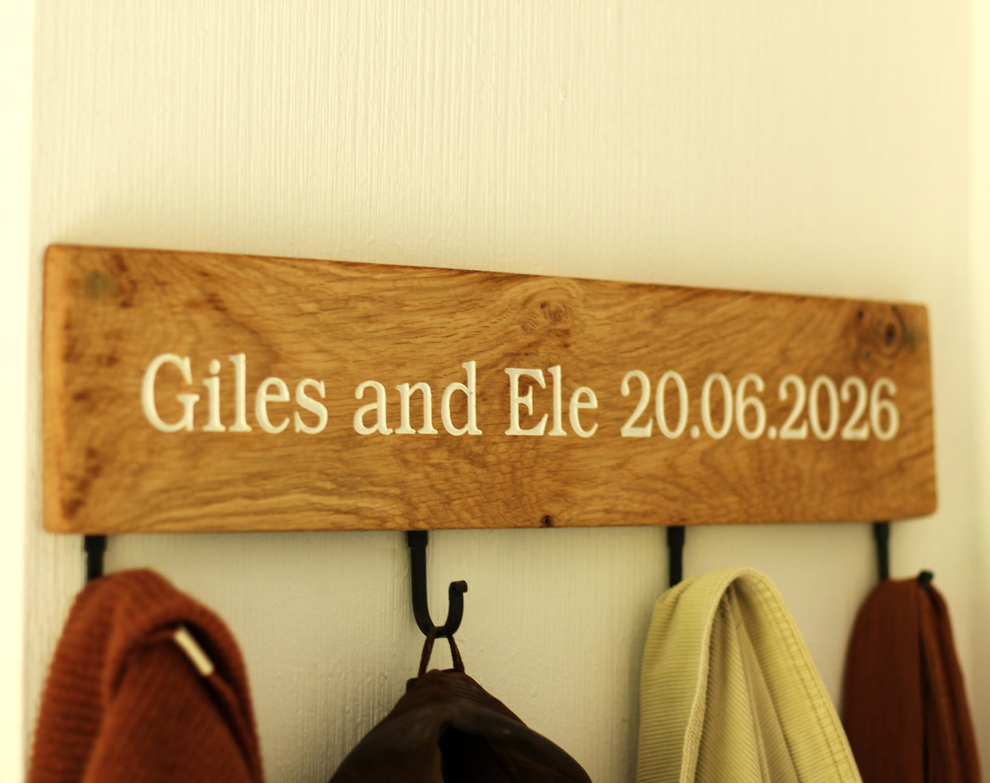 Wooden plaque with 'Giles and Ele 20.06.2026' on a wall with hooks holding coats.