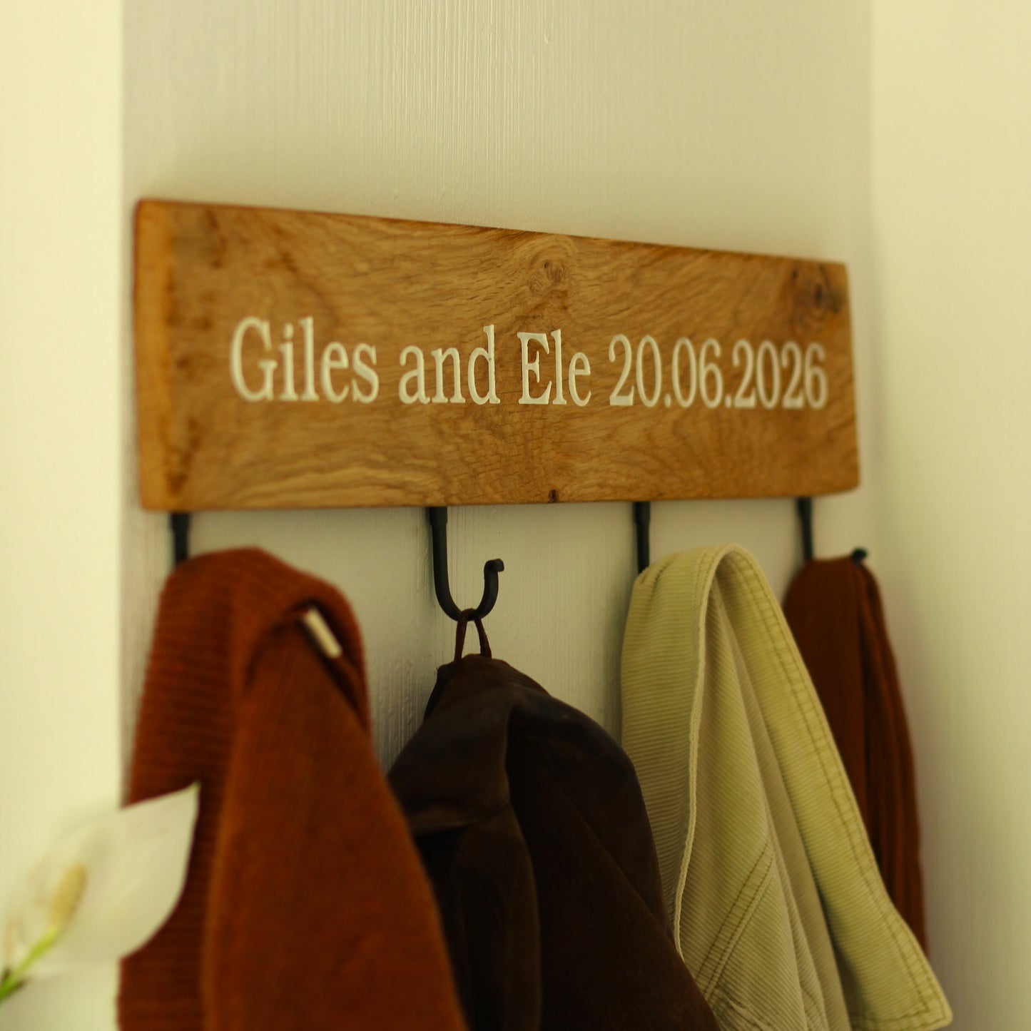 Wooden plaque with 'Giles and Ele 20.06.2026' on a wall with hanging coats.