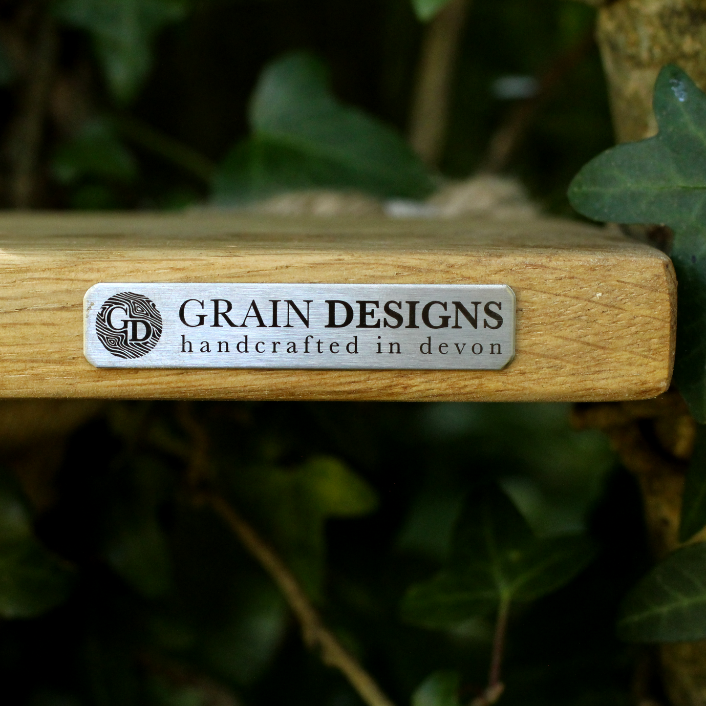 A close-up of a wooden block stamp featuring a silver metal label with the brand name 'GRAIN DESIGNS' and the tagline 'handcrafted in devon', set against a blurred background of green ivy leaves.