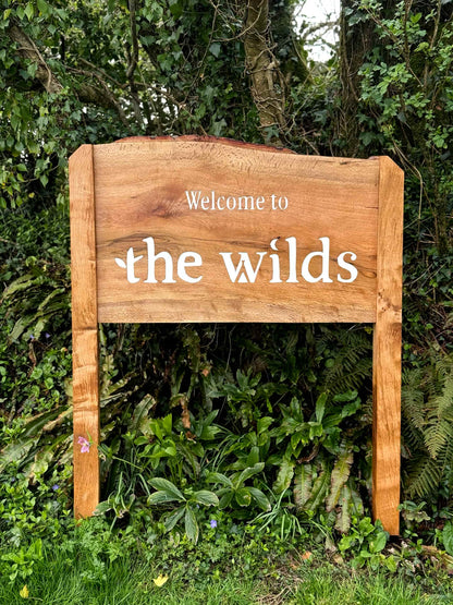 A rustic wooden sign with the words 'Welcome to the wilds' in white lettering, set against a lush green garden background.
