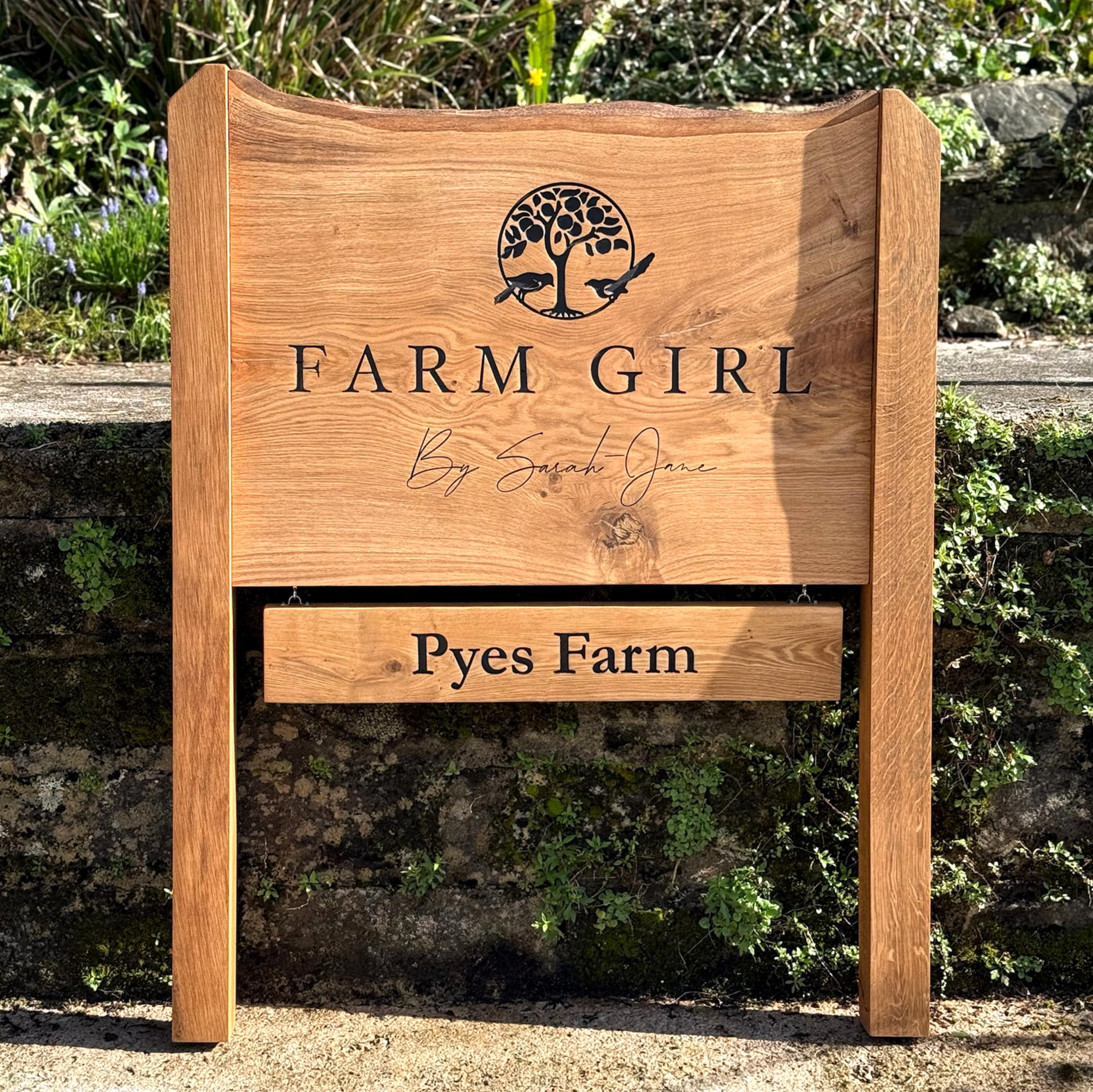 A rustic wooden sign for 'Farm Girl By Sarah Jones' and 'Pyes Farm' displayed outdoors against a stone wall with green foliage.