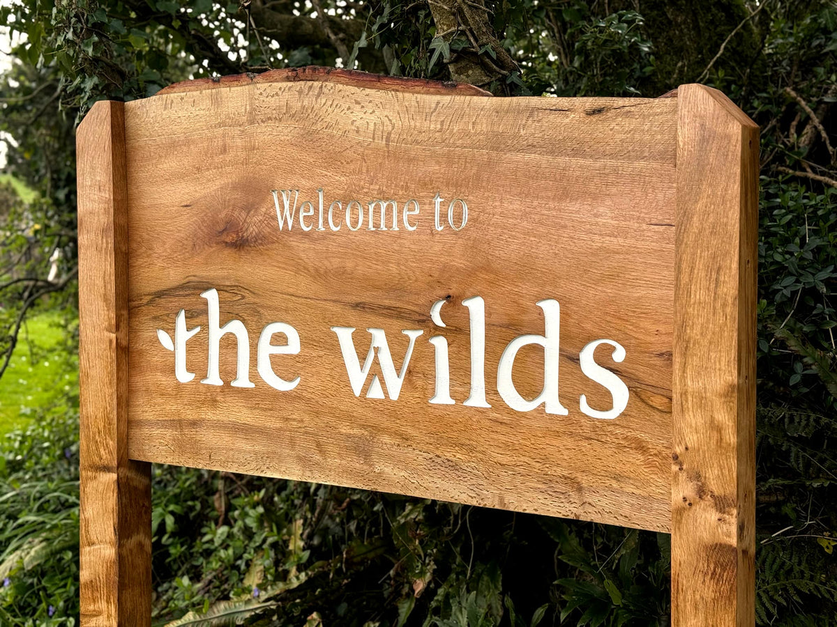 A rustic wooden sign with the words 'Welcome to the wilds' in white lettering, set against a background of green foliage.