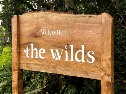 A rustic wooden sign with the words 'Welcome to the wilds' in white lettering, set against a background of green foliage.