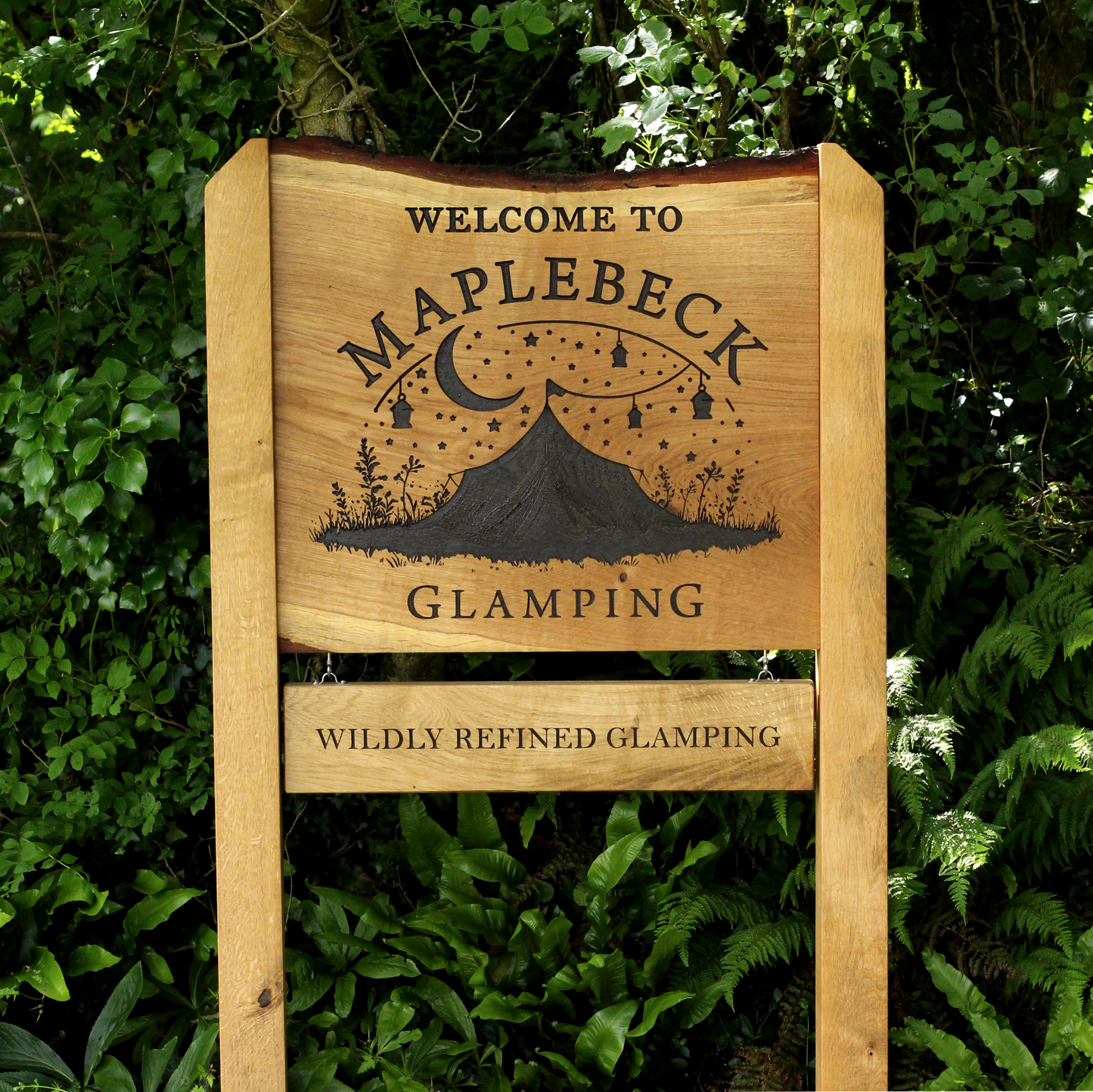 A rustic wooden sign reading 'WELCOME TO MAPLEBECK GLAMPING' with a mountain and moon illustration, mounted outdoors in front of dense green foliage. A smaller hanging plaque below reads 'WILDLY REFINED GLAMPING'.