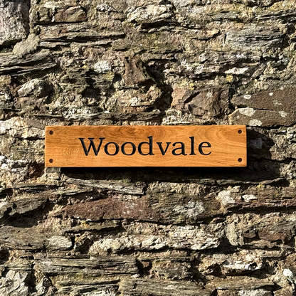 A rectangular wooden sign engraved with the name 'Woodvale' in black lettering, mounted on a rustic stone wall.