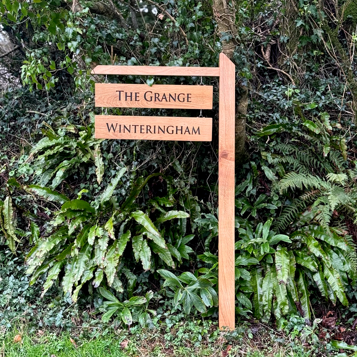 A rustic wooden signpost with two horizontal slats reading 'THE GRANGE' and 'WINTERINGHAM' in black lettering, set against a lush background of green ferns and foliage.