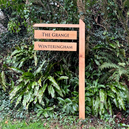 A rustic wooden signpost with two horizontal slats reading 'THE GRANGE' and 'WINTERINGHAM' in black lettering, set against a lush background of green ferns and foliage.