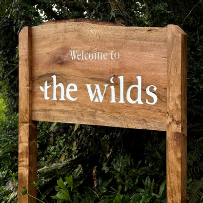 A rustic wooden sign with the words 'Welcome to the wilds' in white lettering, set against a lush green outdoor background of foliage.