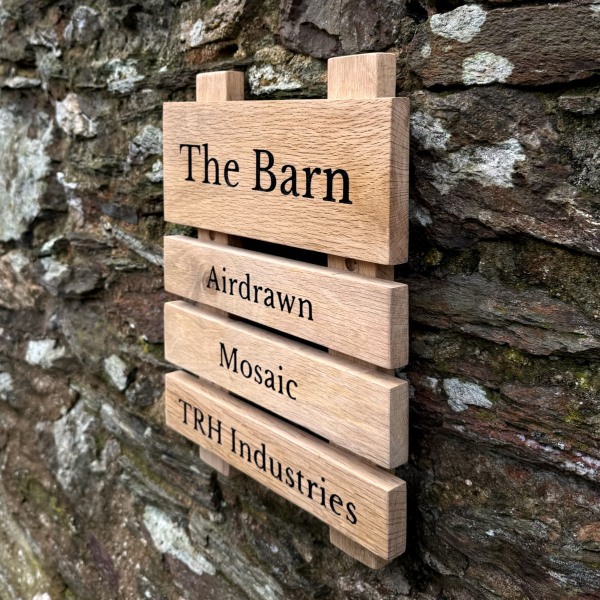 A rustic wooden slat sign reading 'The Barn' with engraved names 'Airdrawn', 'Mosaic', and 'TRH Industries' mounted against a weathered stone wall.