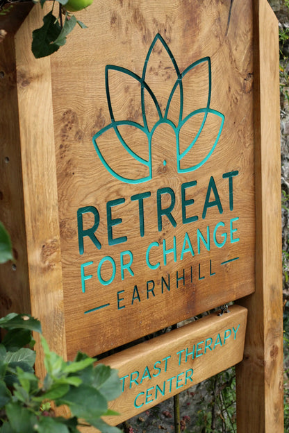 Wooden sign with 'Retreat for Change Earnhill' and 'Trust Therapy Center' text on a natural background.