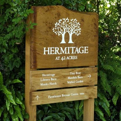 Wooden sign for Hermitage at 42 Acres with tree logo and directional information, surrounded by green foliage.