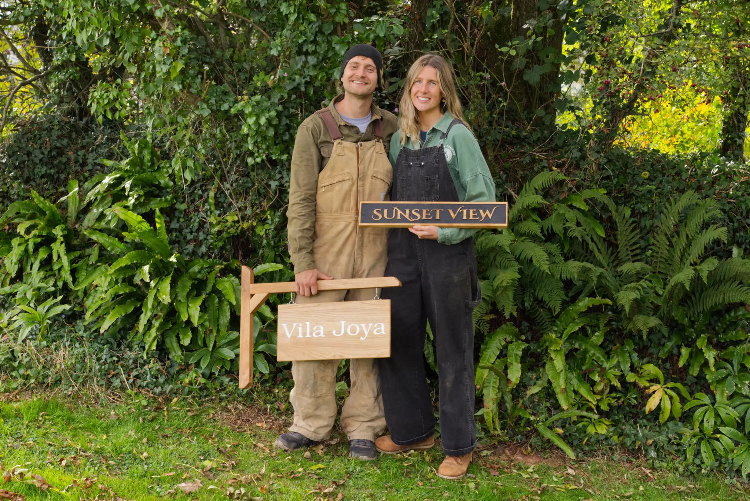 Two people holding wooden signs with text in a garden setting