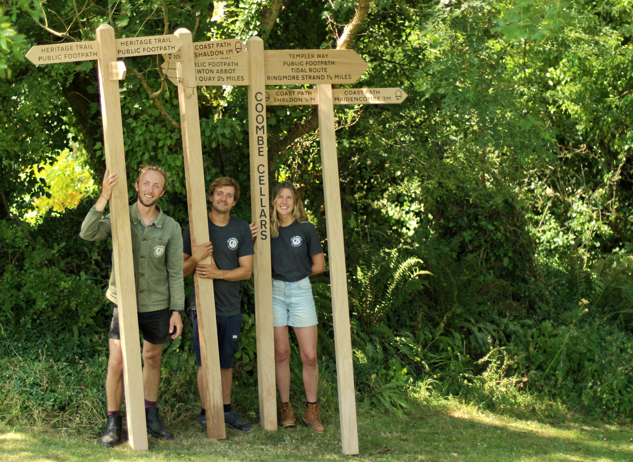 Three people standing outdoors in front of a large wooden directional signpost made of natural wood. The signpost has multiple arrows pointing to different locations, including 'Coastal Path', 'Coombe Cliffs', and others.